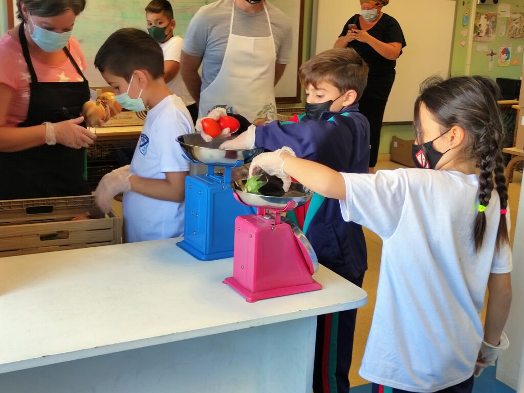Niños y niñas pesan los productos ante la atenta mirada de sus profesores y profesoras.